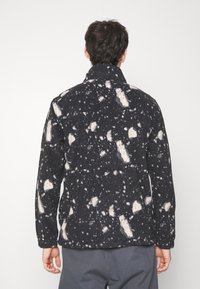 Giacca in pile nera con motivo a macchie bianche, dotata di collo e maniche lunghe, vista da dietro su uno sfondo bianco.