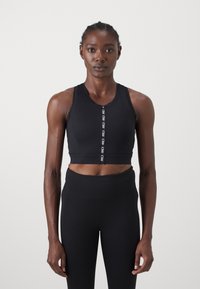 Soutien desportivo preto com fecho à frente, feito de tecido suave. Design ajustado com uma bainha canelada e faixa com logótipo. Usado com leggings pretas de cintura alta.