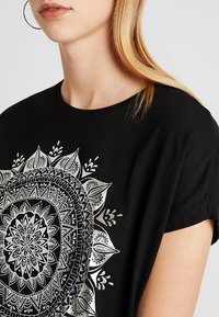 Svart bomull T-shirt med en central silvermandala-design som har intrikata mönster och bladaccenter. Korta ärmar och en avslappnad passform.