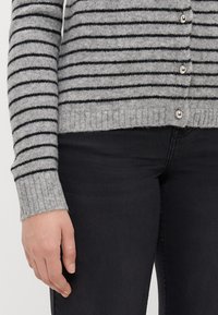 Gråstripet cardigan laget av myk stoff med svarte horisontale linjer, med sølvknapper og ribbestrikkede mansjetter, stylet med mørke jeans.