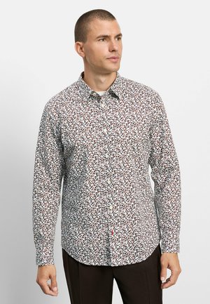 Uomo che indossa una camicia bianca a maniche lunghe con un piccolo motivo floreale marrone e nero, in piedi davanti a uno sfondo chiaro e uniforme.