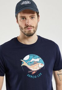 T-shirt marine avec un graphique de poisson en couleurs vives. Tissu doux, col rond et manches courtes. "ARMOR LUX" imprimé en dessous du design.