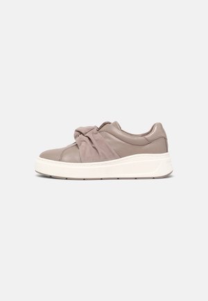 Taupe slip-on sneaker med stor stofsløjfe på toppen og tyk hvid sål, vist fra siden mod en ensfarvet baggrund.