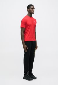 Maglietta di cotone rossa a maniche corte con piccolo logo, abbinata a pantaloni della tuta neri con dettaglio stampato in oro e scarpe da ginnastica nere.