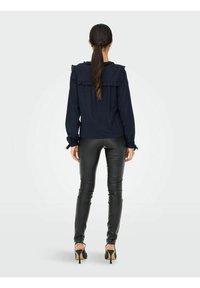 Blouse bleu marine avec des accents froncés sur les épaules et les poignets, associée à un pantalon en cuir noir brillant et des chaussures à talons.