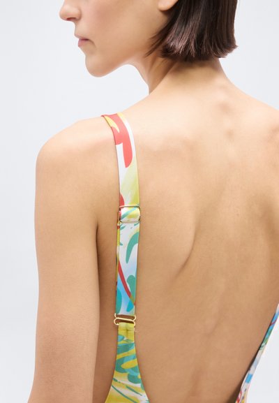 Γυναίκα με κοντά μαλλιά που φοράει πολύχρωμο μαγιό με ρυθμιζόμενους ιμάντες, εμφανίζεται από πίσω σε απλό φωτεινό φόντο.