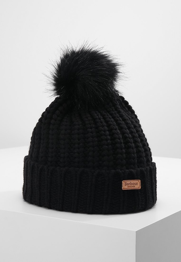 Bonnet noir en tricot épais avec un gros pompon duveteux sur le dessus et une petite étiquette en cuir marron portant l'inscription « Barbour England » sur le revers.