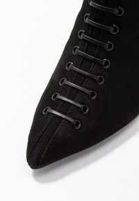 Bottine en daim noir à bout pointu, avec six œillets métalliques et lacets noirs sur le devant pour la fermeture. Texture lisse.