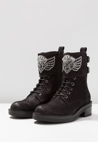 Botas negras hasta el tobillo hechas de cuero suave, con un ala decorativa de tachuelas en la parte superior, cordones y una cremallera lateral. Tacón grueso.