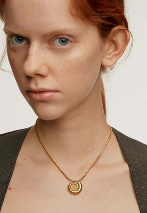 Collar de oro con un colgante en espiral, con un acabado suave. Diseño de cadena delgada sobre una parte superior gris, resaltando los detalles del colgante.