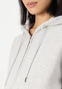 Ljusgrå zip-hoodie med fodrad huva, dragsko och metalliska detaljer. Har mjukt, texturerat tyg för komfort.