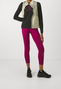 Idrottare bär en beige zip-väst över en svart långärmad tröja och livfullt rosa leggings. Svarta löparskor fulländar outfiten.