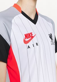 Biały trykot w paski z czerwonymi akcentami, czarnymi rękawami, dekoltem w serek oraz logo Nike Air w kolorze czerwonym. Na piersi znajduje się godło Liverpool FC.