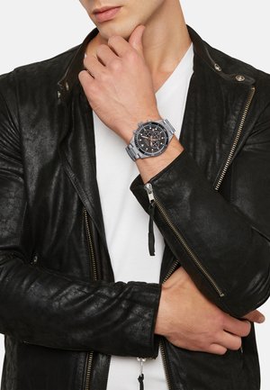 Sølv chronograph ur med sort skive, sølv detaljer og en rustfri stålrem, båret på et håndled mod en sort læderjakke.