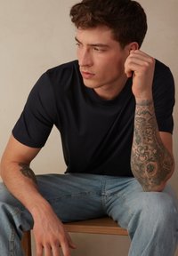 Námořnické triko s krátkým rukávem, bavlněná látka, kulatý výstřih. Model s tetováním v modrých džínách, sedící na dřevěné židli.