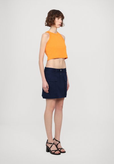 Oransje kort tanktopp, marineblå denimskjørt med frontlommer, og svarte sandaletter med hæl og stropper. Enkel, moderne fasong og rene linjer.