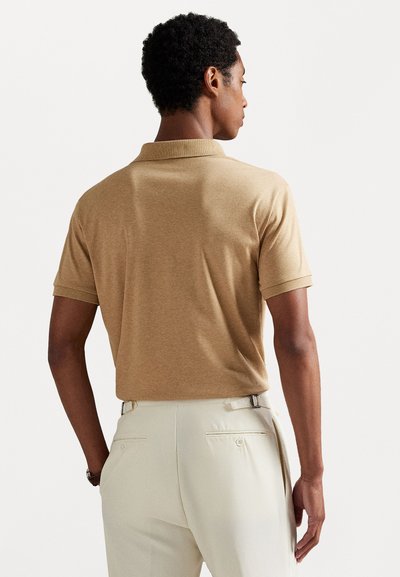 Homem de costas, vestido com uma polo de manga curta castanho claro e calças off-white com ajustes laterais e bolsos traseiros com botão.