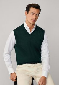 Ciemnozielony sweter bez rękawów w serek, noszony na białej koszuli. Tekstura wydaje się gładka, bez widocznych wzorów.