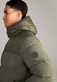 Daunenjacke in dunkelolivgrün mit hohem Kragen, glänzender Oberfläche, gesteppten Panele und einem runden Logo-Patch am Ärmel.
