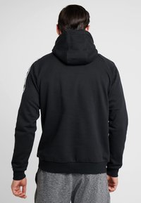 Sudadera negra con una capucha grande, puños y dobladillo acanalados. Presenta rayas blancas a lo largo de los hombros. El material parece suave y con textura.