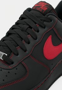 Black leather sneaker with red accents and stitching. Features a rounded toe, flat laces, and a circular logo on the side.