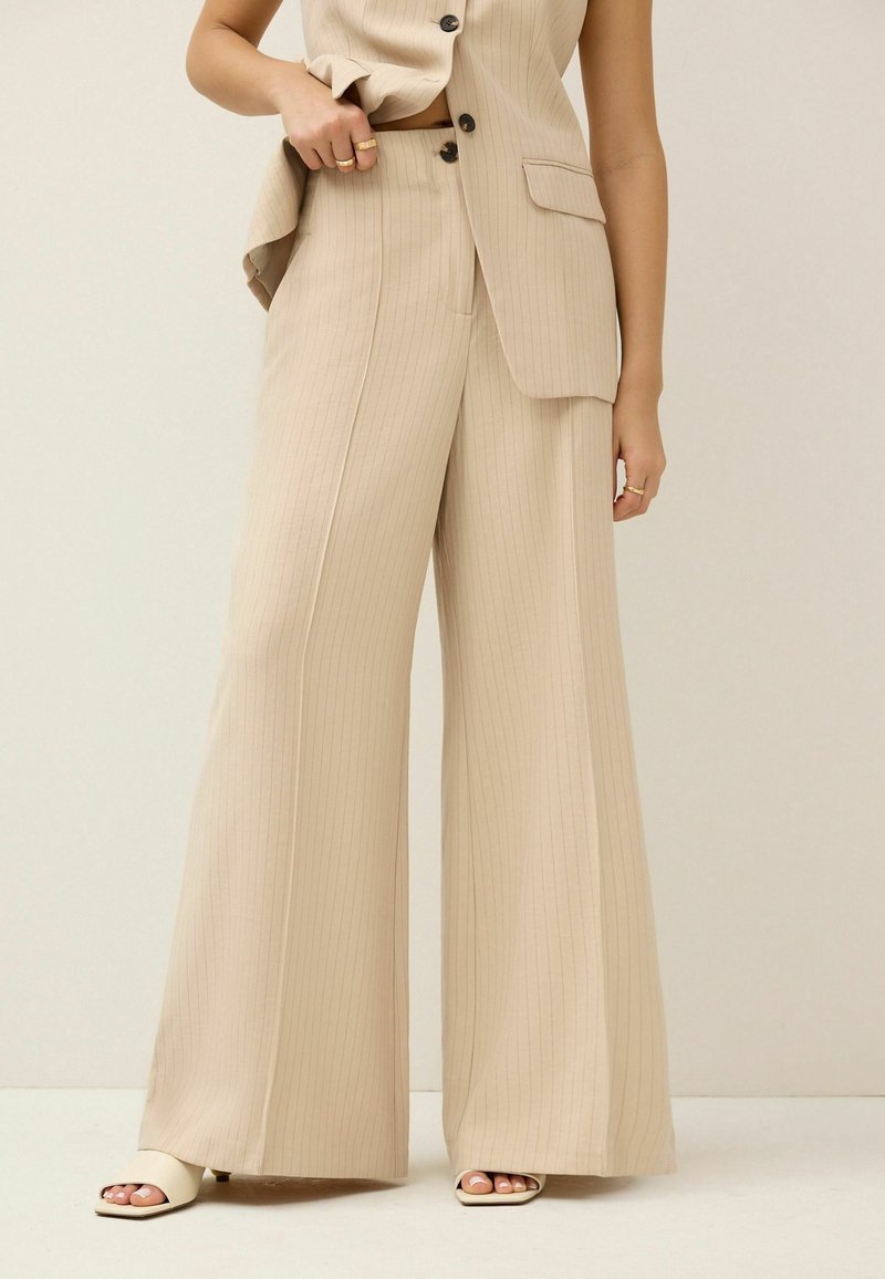 Pantalon beige à fines rayures, jambes larges, en tissu lisse, avec une taille cintrée et une coupe décontractée, associé à des sandales de couleur claire.