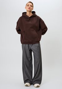 Túlméretes barna kapucnis pulóver kenguruzsebbel, szürke csíkozott bőszárú nadrággal és bézs sportcipővel párosítva. Lágy szövettextúrák láthatók.