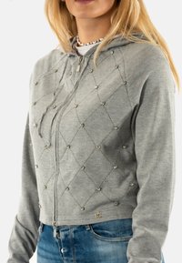 Haut à capuche zippé gris avec un motif en diamant, orné de petites perles en argent. Comprend une capuche avec cordon de serrage et des poignets côtelés.