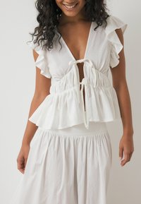 Witte, geplooide blouse met een diepe V-hals en verstelbare strik aan de voorkant. Lichtgewicht, vloeiende stof met een gelaagd peplumontwerp.
