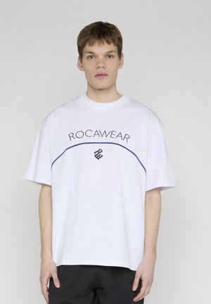 Fiatal férfi rövid barna hajjal, fehér Rocawear pólót és fekete nadrágot visel, egyszerű világos háttér előtt áll.