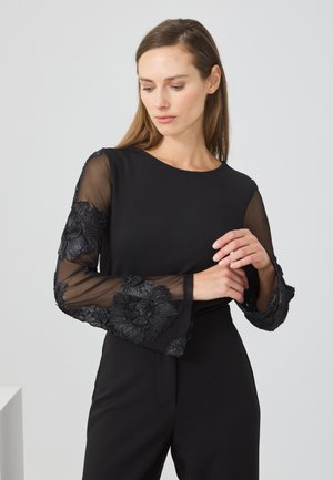 Haut noir avec des manches transparentes ornées de broderies florales complexes. Encolure ronde simple et corsage ajusté. Design élégant et texturé.