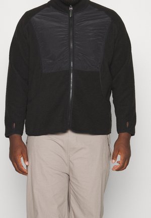 Homme portant une veste polaire noire zippée avec des panneaux en nylon sur la poitrine et un pantalon beige, debout devant un fond uni.