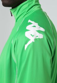 Homme portant un haut zippé vert vif avec un logo de marque blanc représentant deux silhouettes humaines assises dos à dos sur l'épaule.