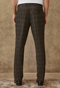 Brown plaid trousers with a straight leg fit. Features two back pockets with buttons and subtle texture. Completed with black shoes.
