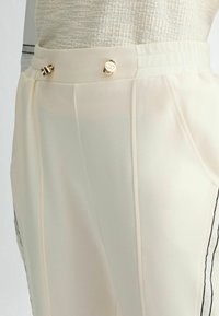 Pantalons blancs en tissu léger et lisse. Présentent des plis et deux boutons dorés au niveau de la taille, ornés de rayures noires.