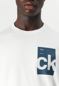 Bijela pamučna majica s tamnoplavim grafičkim etiketom na kojoj piše "Calvin Klein" i "since 1968". Okrugli izrez. Glatka tekstura.