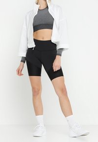 Weiße Netzjacke mit Reißverschluss über einem grauen Crop-Top, kombiniert mit schwarzen Radshorts. Das Modell trägt weiße Sportschuhe und gerippte Knöchelsocken.