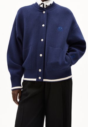 Person iført en mørkeblå cardigan med knapper, hvide kanter og et lille blåt smiley-emblem, kombineret med sorte bukser.