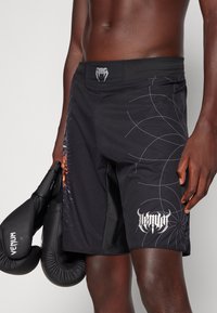 Pantalones cortos de boxeo negros con una cinturilla texturizada, que presentan un diseño gráfico en azul y naranja, y un logo blanco en la parte inferior de la pierna.
