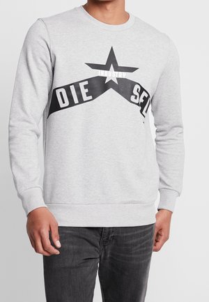 Mężczyzna w jasnoszarej, długorękawowej bluzie z czarną gwiazdą i zakrzywionym napisem "DIESEL INDUSTRY" na piersi, w ciemnoszarych dżinsach.