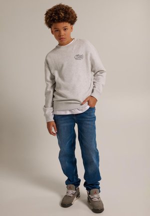 Niño con cabello rizado está de pie vestido con una sudadera gris claro, vaqueros azules y zapatillas grises con logotipos rojos sobre un fondo liso.