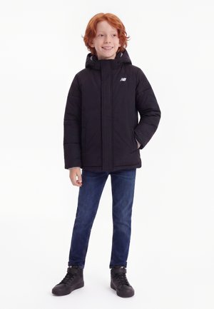 Smilende rødhåret barn iført en sort hættejakke, blå jeans og sorte høje sneakers, stående med den ene hånd i lommen.