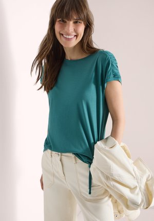 Donna sorridente con capelli castani che indossa una maglia verde acqua con maniche arricciate e pantaloni color crema, tenendo in mano destra una giacca chiara.