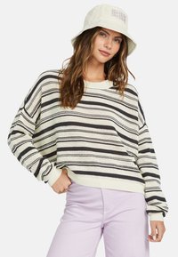 Striped sweater in cream with black and gray stripes. Loose fit, ribbed cuffs, and hem. Paired with light purple pants and a white bucket hat.