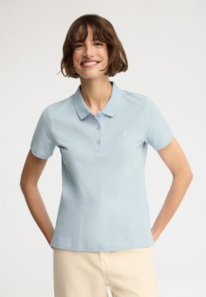 Kobieta uśmiechająca się, ubrana w jasnoniebieską koszulkę polo z krótkim rękawem i beżowe spodnie, stojąca z rękami za plecami na gładkim tle.