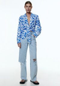 Blaues Blumendruckhemd mit geknoteter Front, kombiniert mit hellblauen, verwaschenen Schlaghosen und schwarzen Sandalen. Glattes Material, lockere Passform.
