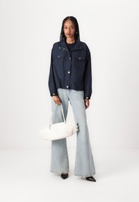 Giacca cropped in nylon blu navy con tasche frontali, bottoni bianchi, abbinata a jeans a vita alta blu chiaro a zampa d'elefante e una borsa bianca.