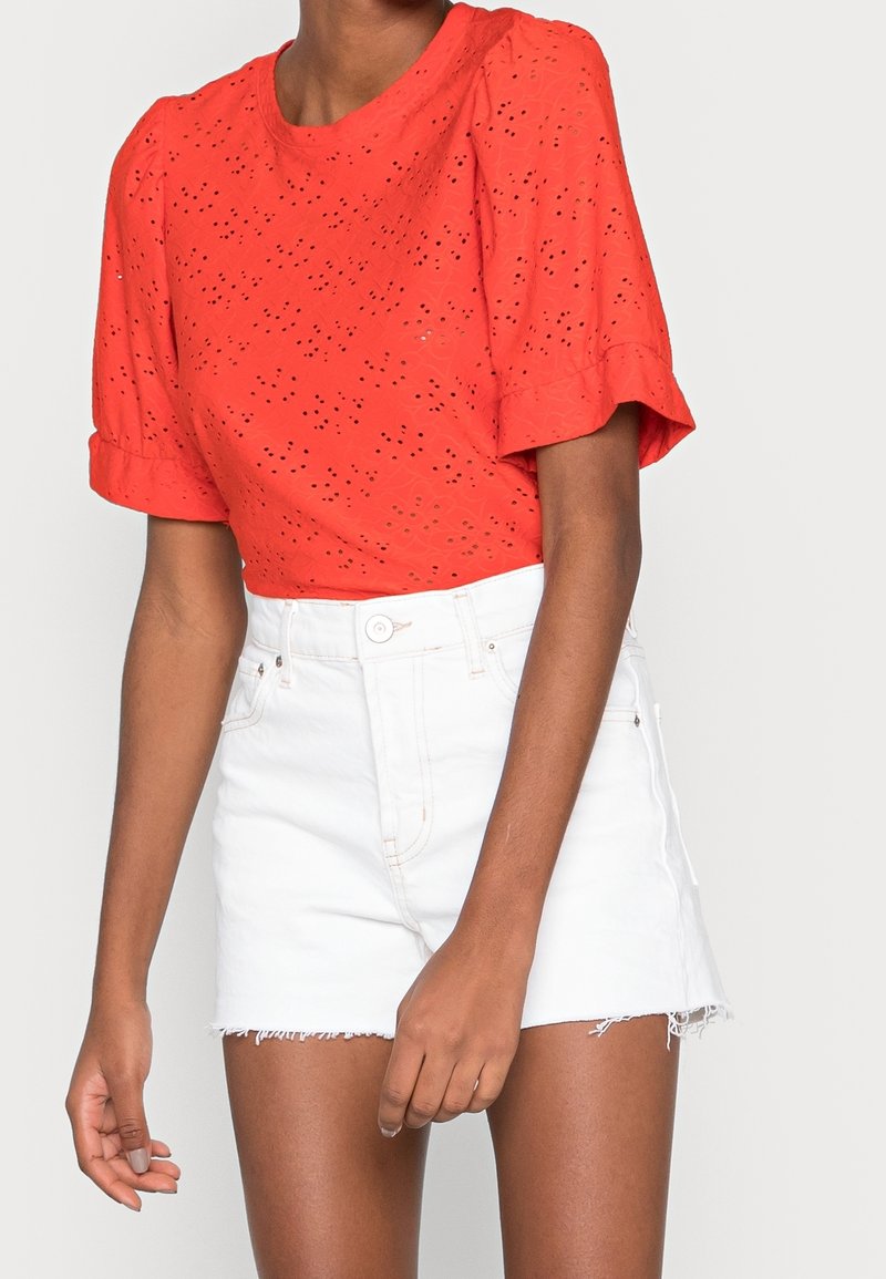 Blouse rouge à œillets avec des manches bouffantes et un col rond, associée à un short en denim blanc déchiré avec un ourlet effiloché.