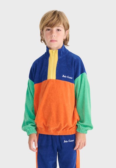 Enfant aux cheveux châtain clair portant un survêtement coloré avec des sections bleues, orange, vertes et jaunes, debout devant un fond clair uni.
