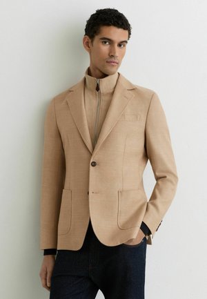 Hombre vistiendo un blazer beige sobre una chaqueta marrón con cremallera y pantalones oscuros, de pie contra una pared blanca lisa con una mano en el bolsillo.